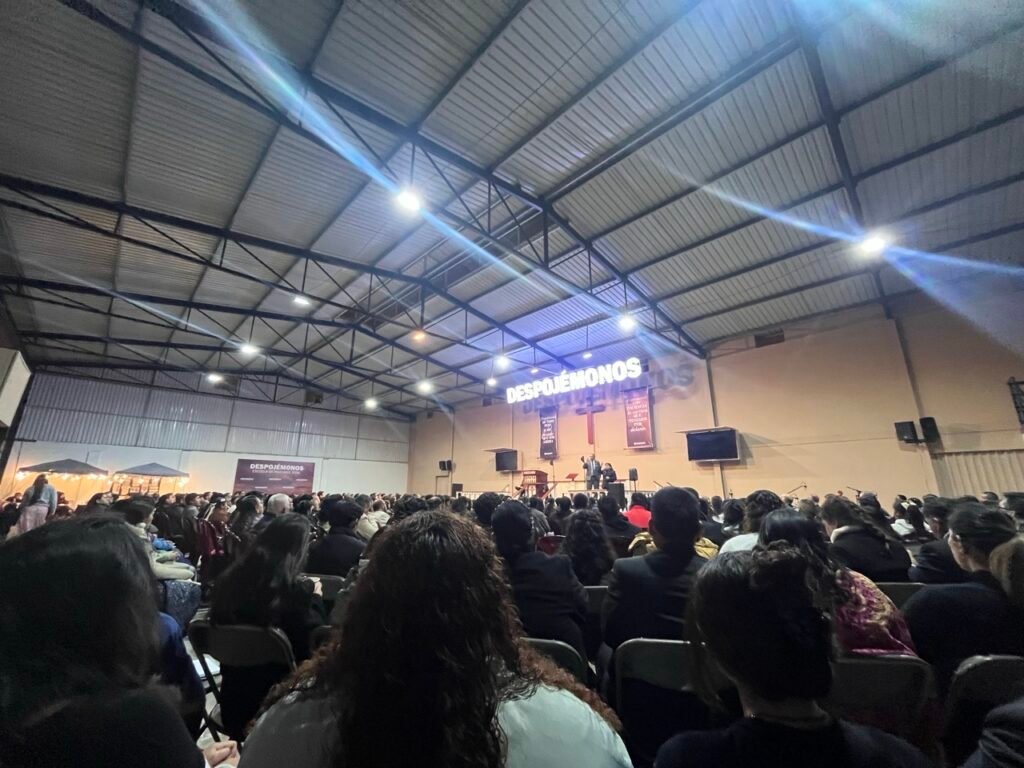 A large congregation sits in an auditorium listening to a message, with a brightly lit sign reading "DESPOJEMONOS" suspended above the speaker.